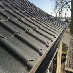 Nowy, ciemnoszary dach z blachodachówki, z zamontowaną rynną, widok z rusztowania na tle błękitnego nieba i drzew