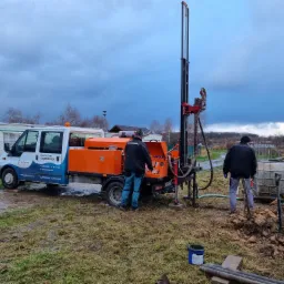 Wiertnica zamontowana na pomarańczowo-białym samochodzie dostawczym podczas wiercenia studni; dwóch pracowników w roboczych ubraniach nadzoruje proces, w tle przyczepa kempingowa i pochmurne niebo.