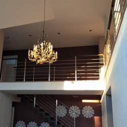 Kristal - Dwupoziomowe wnętrze domu z metalowymi schodami i balustradą, oświetlone ozdobnym żyrandolem.