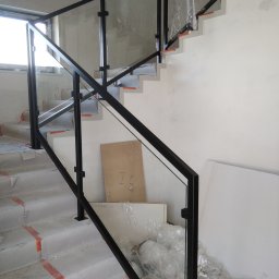 Kristal - Montaż szklanej balustrady z czarnymi, metalowymi słupkami na betonowych schodach w trakcie budowy, zabezpieczonych taśmą. Widoczne materiały budowlane i folie ochronne.