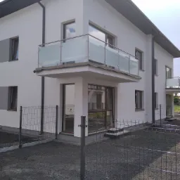 Nowoczesny budynek z balkonami zabezpieczonymi szklanymi balustradami ze stalowymi poręczami, widok z zewnątrz przez metalowe ogrodzenie.