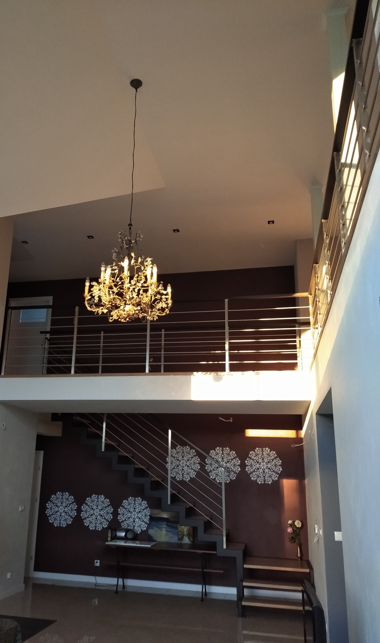 Dwupoziomowe wnętrze domu z metalowymi schodami i balustradą, oświetlone ozdobnym żyrandolem.