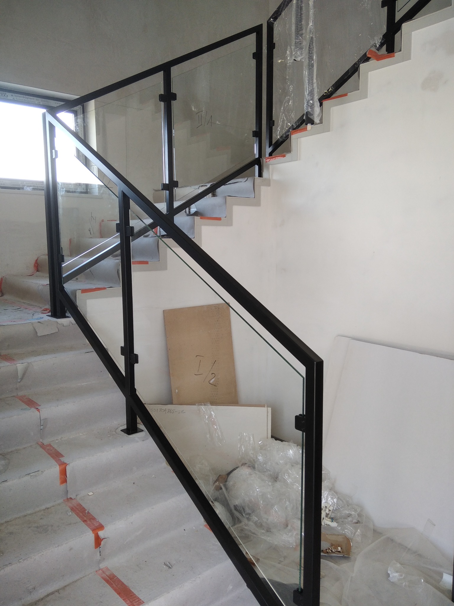 Montaż szklanej balustrady z czarnymi, metalowymi słupkami na betonowych schodach w trakcie budowy, zabezpieczonych taśmą. Widoczne materiały budowlane i folie ochronne.