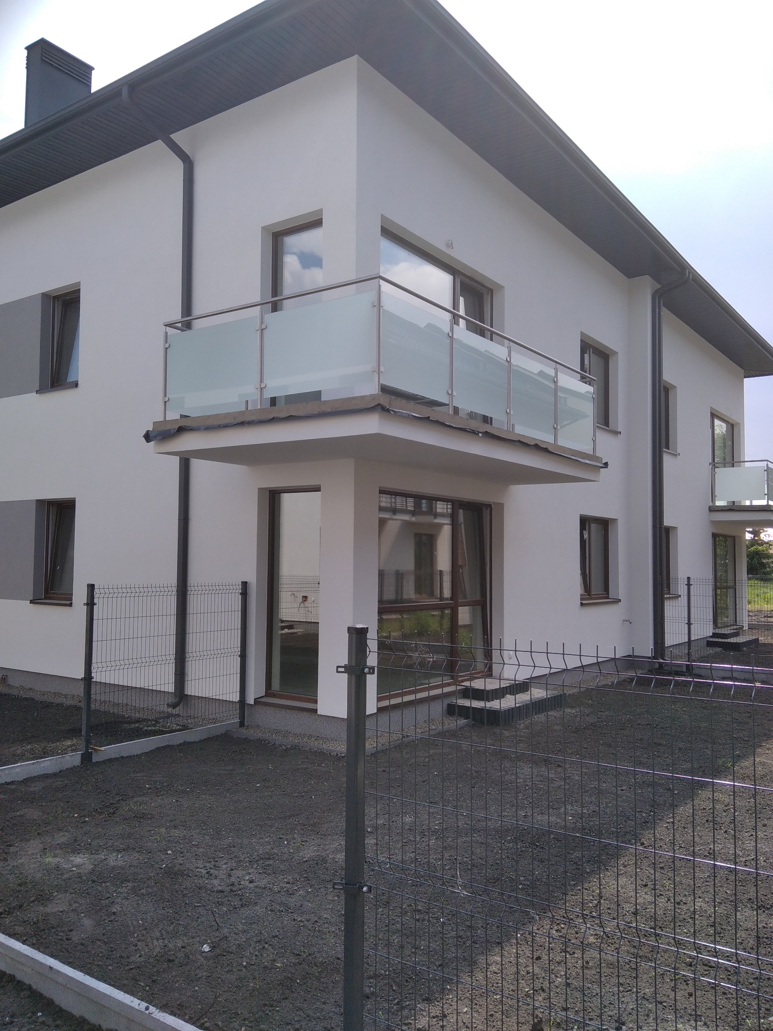 Nowoczesny budynek z balkonami zabezpieczonymi szklanymi balustradami ze stalowymi poręczami, widok z zewnątrz przez metalowe ogrodzenie.