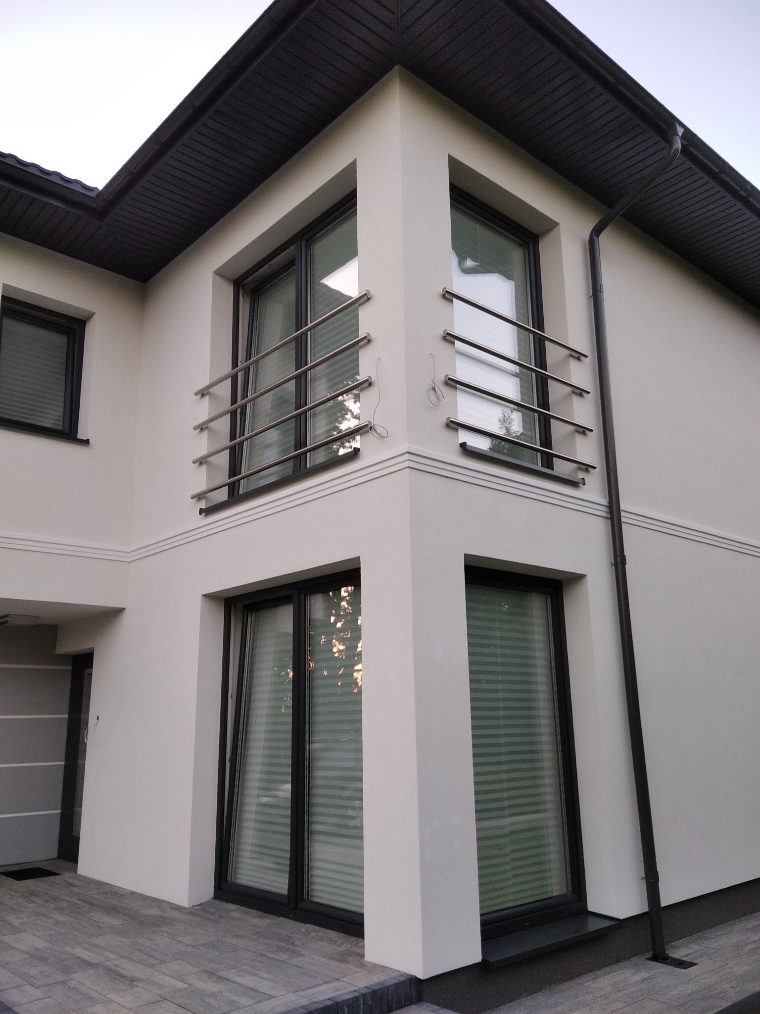 Nowoczesny budynek z balkonami zabezpieczonymi poziomymi, metalowymi prętami, widok z dołu na narożną część budynku, ciemna stolarka okienna i orynnowanie.