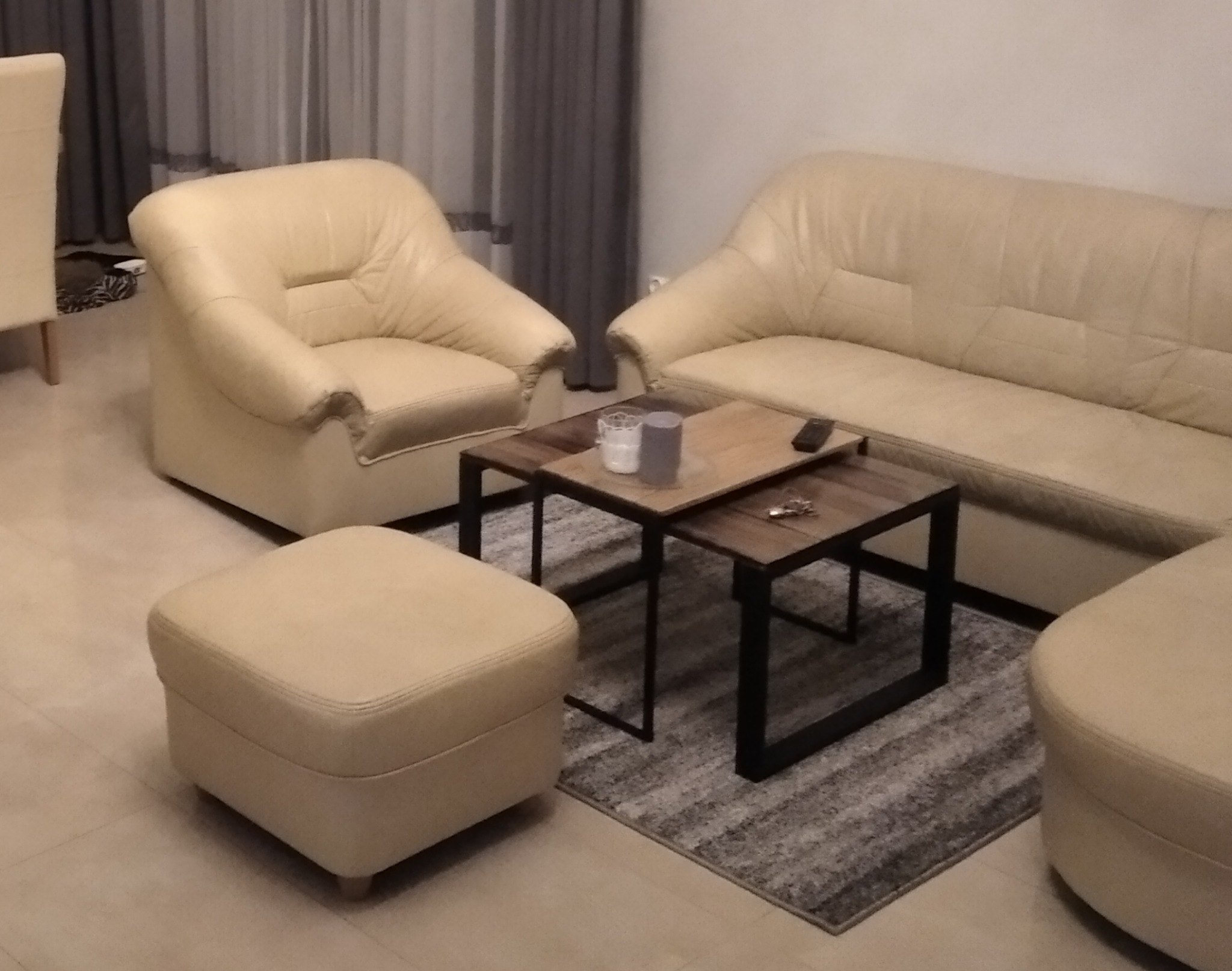 Jasna skórzana sofa i fotel z podnóżkiem, stolik kawowy z dwoma blatami na metalowych nogach, szary dywan w tle zasłony.