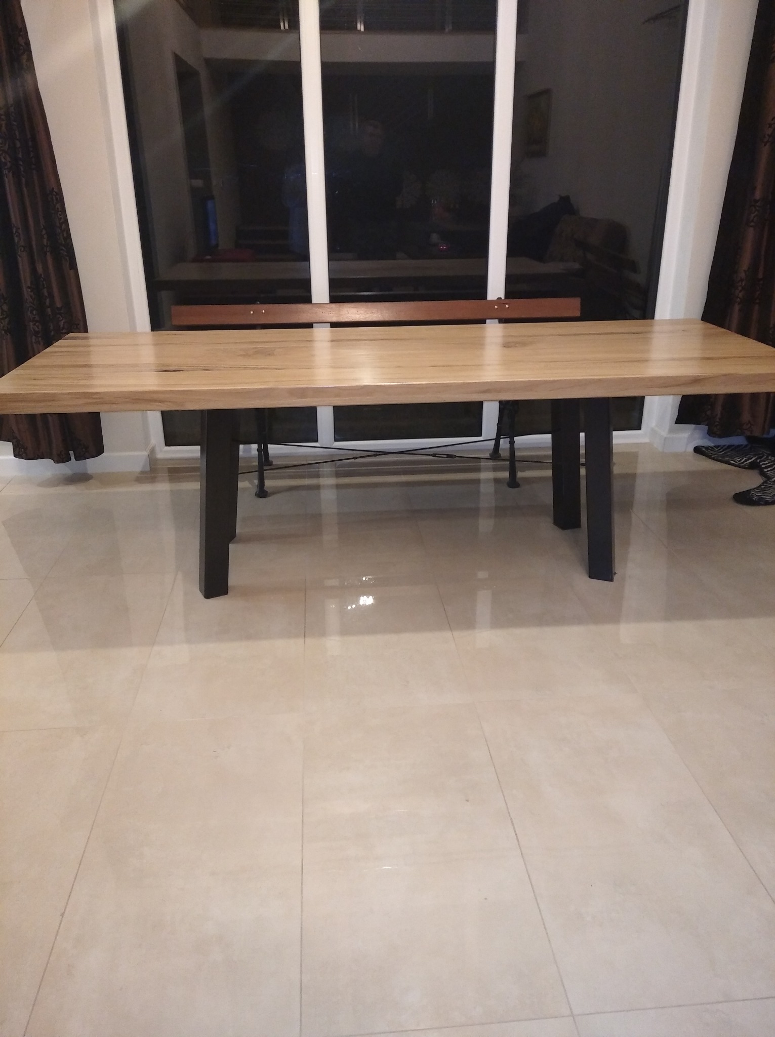 Stół z drewnianym blatem i czarnymi, metalowymi nogami, ustawiony na jasnych płytkach podłogowych, z widokiem na okno w tle.