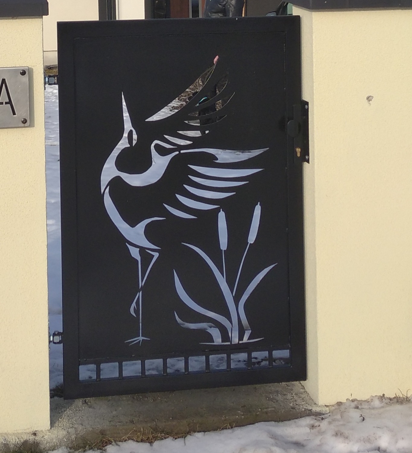 Czarna metalowa furtka z laserowo wycinanym motywem żurawia i roślinności wodnej, zamontowana przy jasnej ścianie budynku.