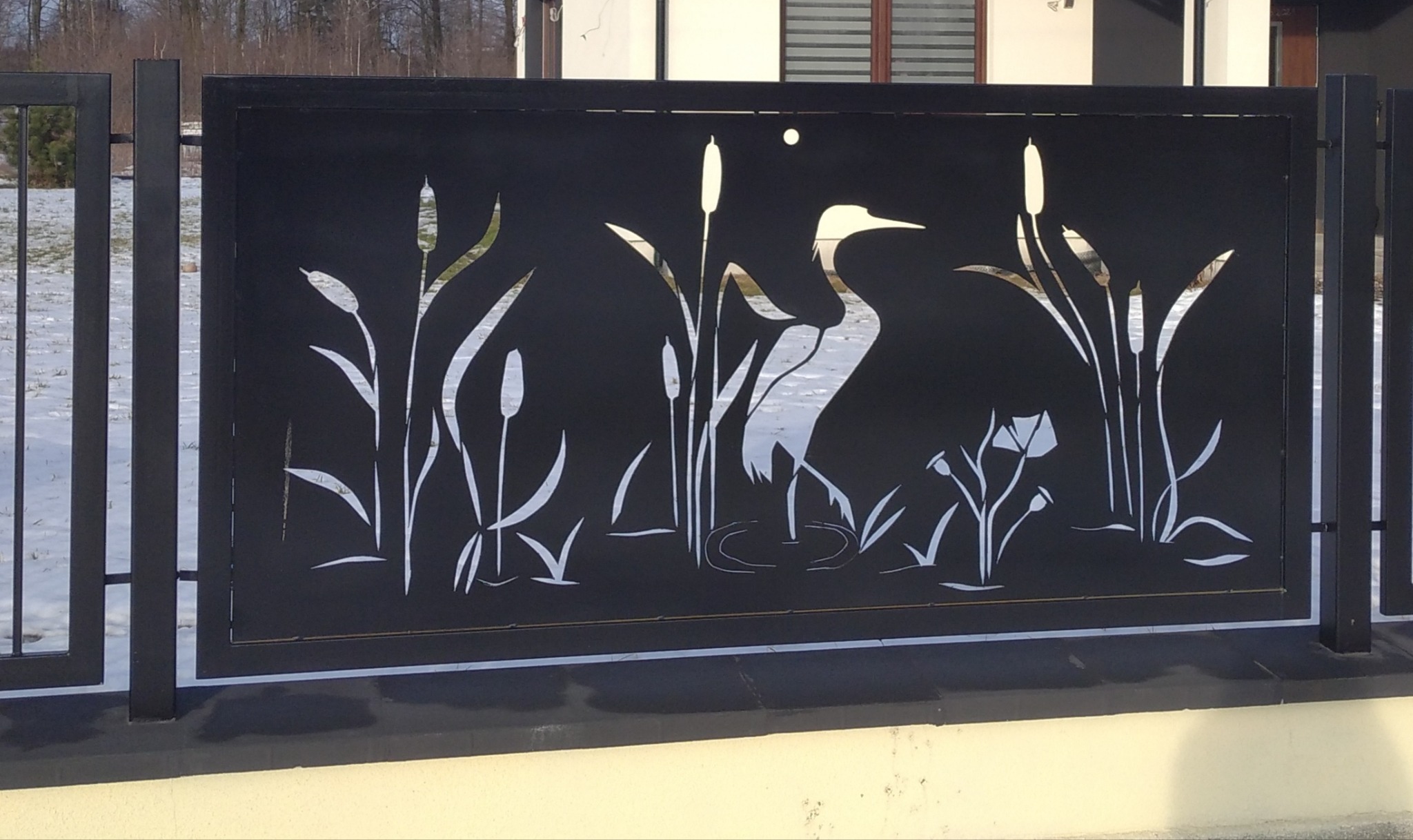 Panel ogrodzeniowy z czarnego metalu, dekorowany wycinanym laserowo motywem roślinnym i wizerunkiem czapli, w tle zasypany śniegiem ogród.