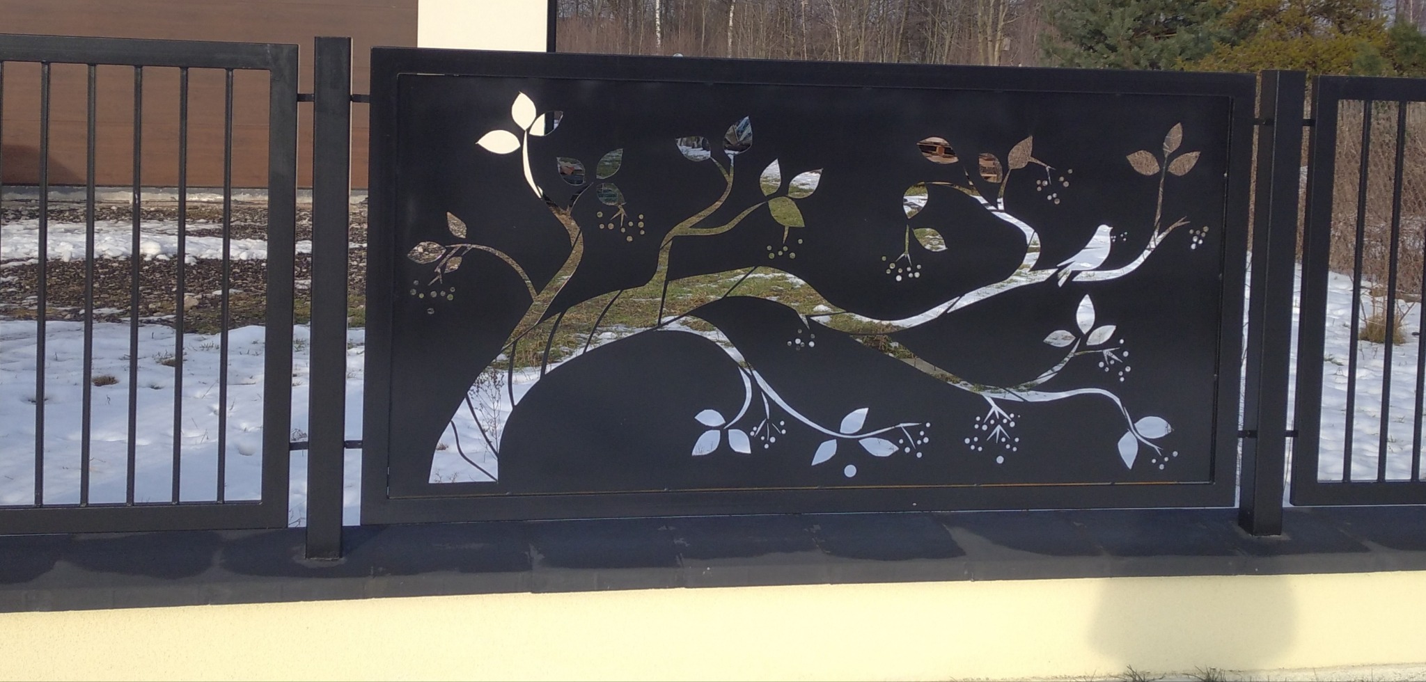 Czarny metalowy panel ogrodzeniowy z wycinanym laserowo motywem drzewa z ptakiem, część ogrodzenia posesji w zimowej scenerii.