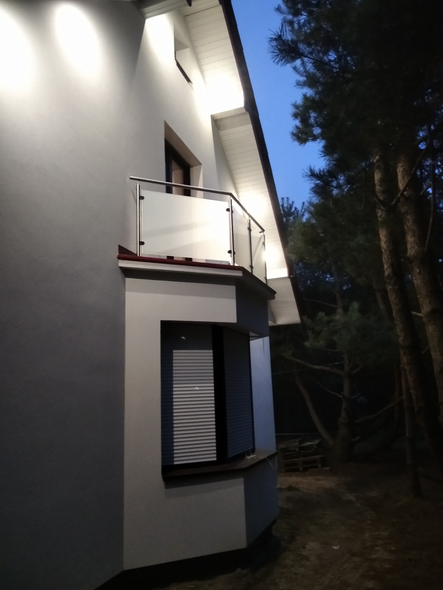 Nowoczesny balkon z szklaną balustradą i metalową poręczą, oświetlony od góry, na tle wieczornego nieba i drzew