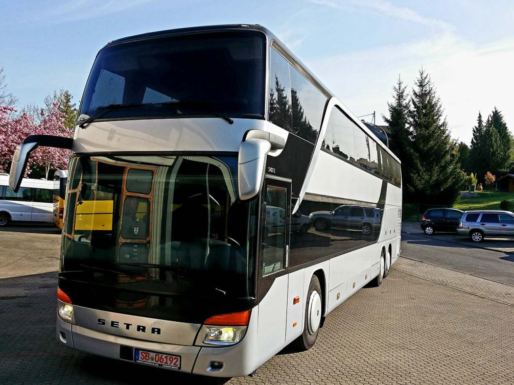 Dwupokładowy autokar Setra w kolorze srebrno-czarnym, zaparkowany na brukowanym placu, z odbiciami samochodów i drzew w oknach, tablica rejestracyjna SB 06192.