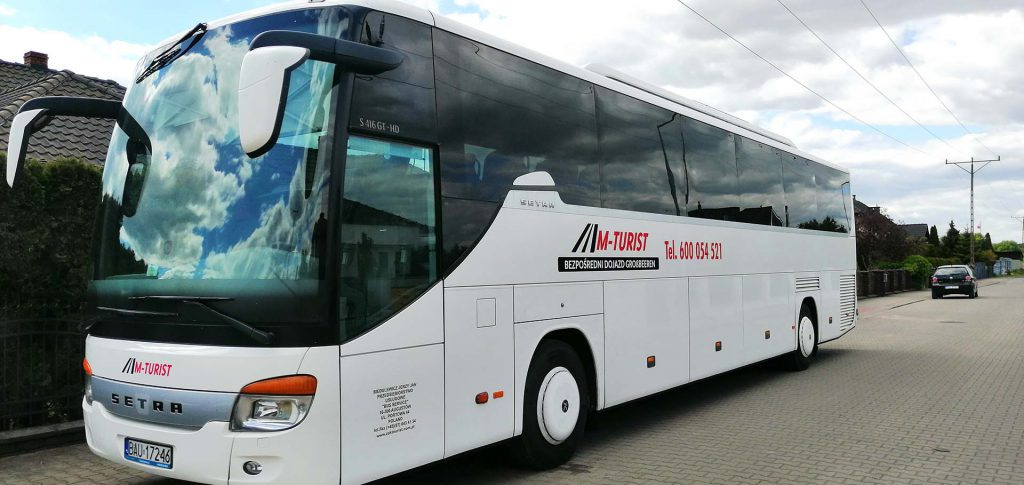 Biały autokar marki Setra z logo firmy M-Turist na tle brukowanej drogi i pochmurnego nieba, z odbiciem chmur w szybach.