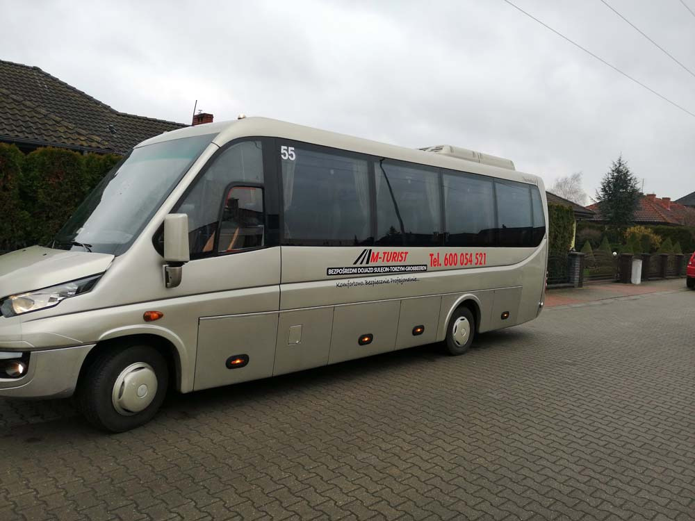 Srebrny bus turystyczny z numerem 55 i logo firmy M-Turist parkuje na brukowanej ulicy przed domami.