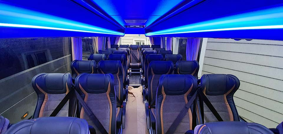 Wnętrze autobusu turystycznego z rzędami foteli w kolorze granatowym i szarym, oświetlenie LED w kolorze niebieskim, widok z perspektywy pasażera.