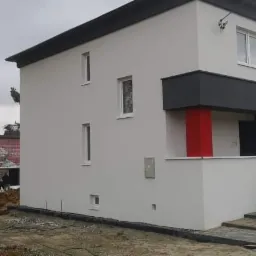 Dwukondygnacyjny dom z płaskim dachem i elementami w kolorze szarym i czerwonym, widoczny w trakcie prac wykończeniowych elewacji zewnętrznej.