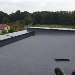 Szary, płaski dach pokryty papą termozgrzewalną z widocznymi obróbkami blacharskimi, narzędziami i fragmentem buta dekarza na tle zabudowań i drzew.