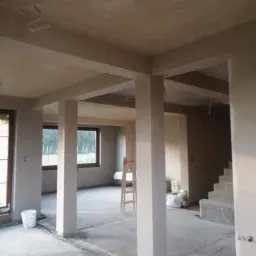 Wnętrze domu w trakcie prac wykończeniowych, widoczne kolumny i ściany pokryte świeżym tynkiem, częściowo poprowadzone instalacje elektryczne i schody w surowym stanie.