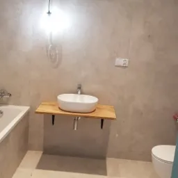 Nowoczesna łazienka z owalną umywalką nablatową na drewnianym blacie, wiszącą lampą z pojedynczą żarówką i szarą, teksturowaną ścianą.