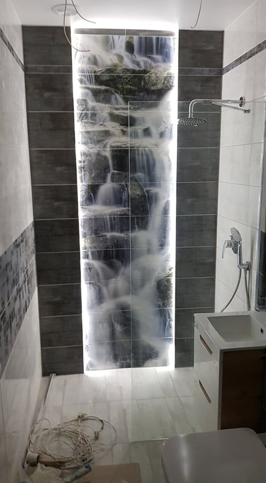 Wykończona łazienka z podświetlanym panelem ściennym imitującym wodospad, szara glazura, biała umywalka z szafką i chromowana armatura.