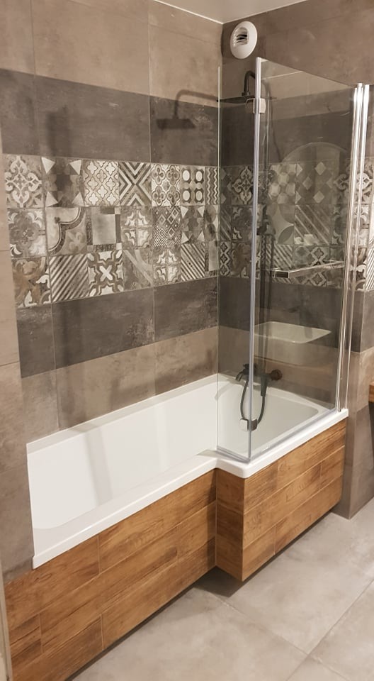 Nowoczesna łazienka z wanną obudowaną drewnopodobnymi płytkami, szklany parawan nawannowy, ściana wyłożona szarymi płytkami z dekoracyjnym pasem mozaiki.