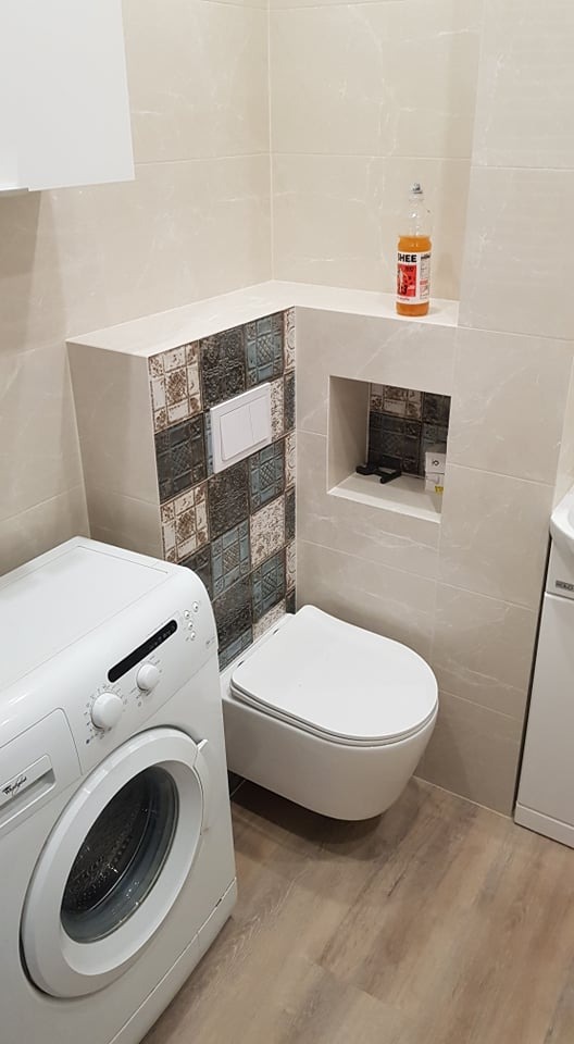 Wykończona łazienka z podwieszaną toaletą, pralką i beżowymi płytkami z dekoracyjnym panelem mozaikowym za spłuczką; na półce w ścianie drobne akcesoria, a na górnej półce butelka płynu.