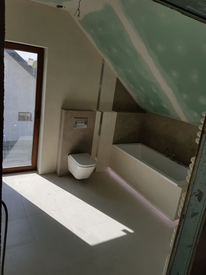 Poddasze łazienki w trakcie wykańczania: widoczna wanna z podświetleniem LED, podwieszana toaleta, płytki na ścianach i podłodze, oraz sufit z płyt gipsowo-kartonowych przed szpachlowaniem,...