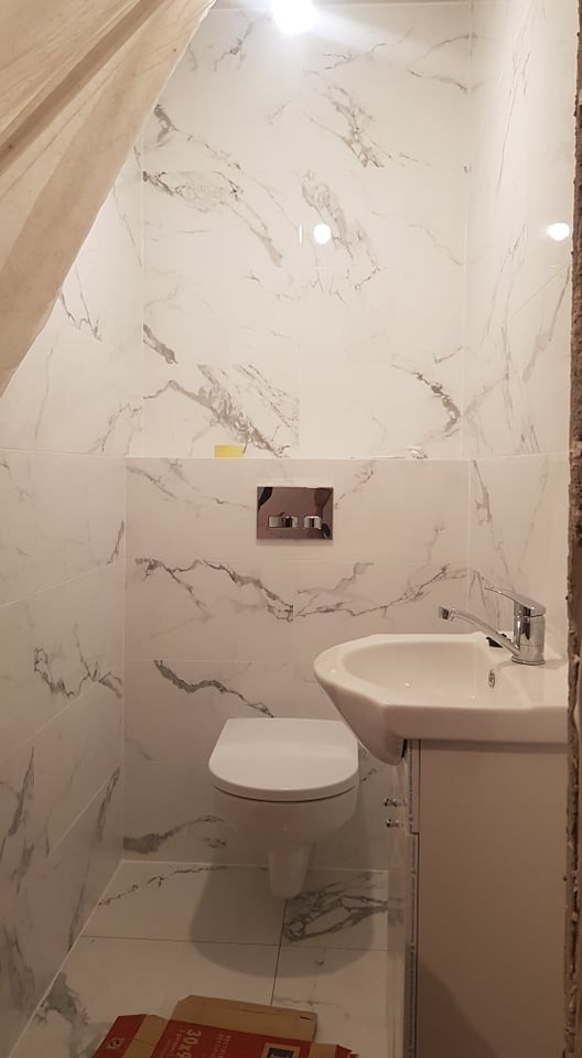 Mała łazienka w trakcie wykańczania z białymi płytkami imitującymi marmur, wisząca toaleta i umywalka z szafką, widoczna kartonowa paczka płytek na podłodze.