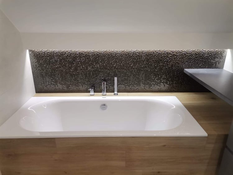 Biała wanna w łazience na poddaszu z oryginalną mozaiką w odcieniach szarości i z oświetleniem LED w niszy nad wanną.