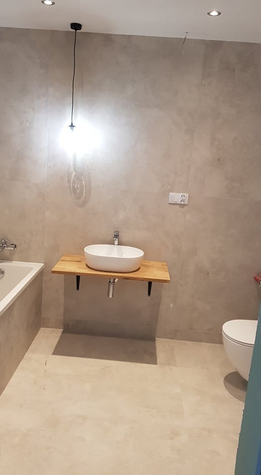 Nowoczesna łazienka z owalną umywalką nablatową na drewnianym blacie, wiszącą lampą z pojedynczą żarówką i szarą, teksturowaną ścianą.