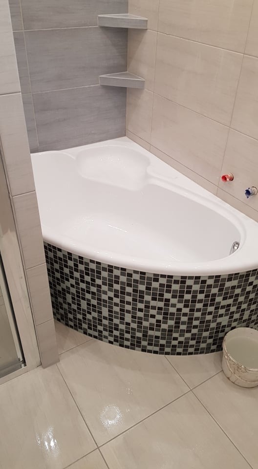 Narożna wanna obudowana mozaiką czarno-białą, z półkami narożnymi w szarym odcieniu i beżowymi płytkami na ścianach, uzupełnona widocznym wiaderkiem z wodą.