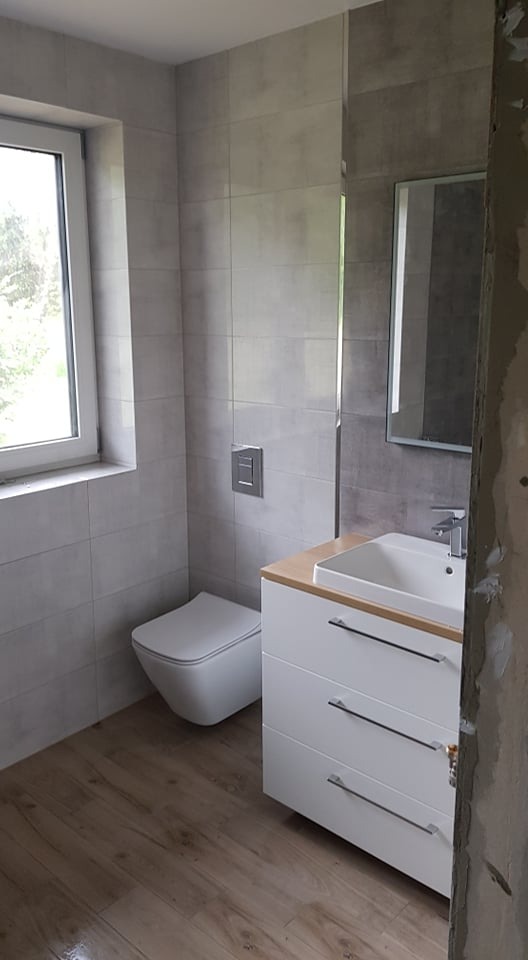 Nowoczesna łazienka z szarymi płytkami, podwieszaną toaletą, białą szafką z umywalką i drewnianym blatem, oraz panelami podłogowymi o wyglądzie drewna. Częściowo wykończone ściany po prawej stronie.