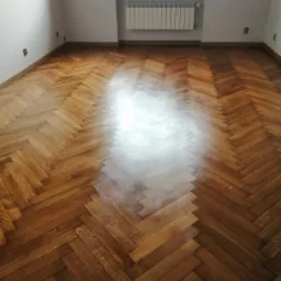 Wycyklinowana podłoga z parkietu ułożonego w jodełkę, widoczne odbicie światła od okna, ściany pomalowane na biało, grzejnik pod oknem.