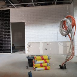 Stanisław Piersa - Wnętrze w trakcie układania płytek: fragment ściany z czarną i białą glazurą, widoczne worki z zaprawą i zwisające kable instalacji elektrycznej.