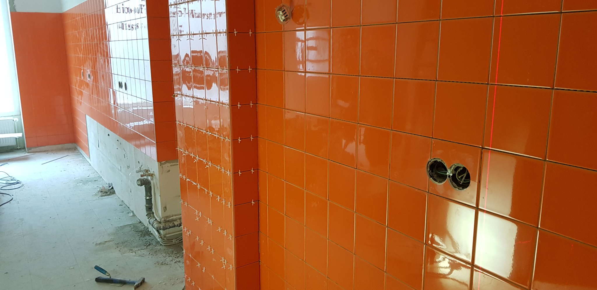 Pomarańczowe, błyszczące płytki ceramiczne na ścianie, ułożone z krzyżykami dystansowymi, widoczne otwory na gniazdka elektryczne i ślady po laserowym wyznaczaniu linii, w tle surowy stan...