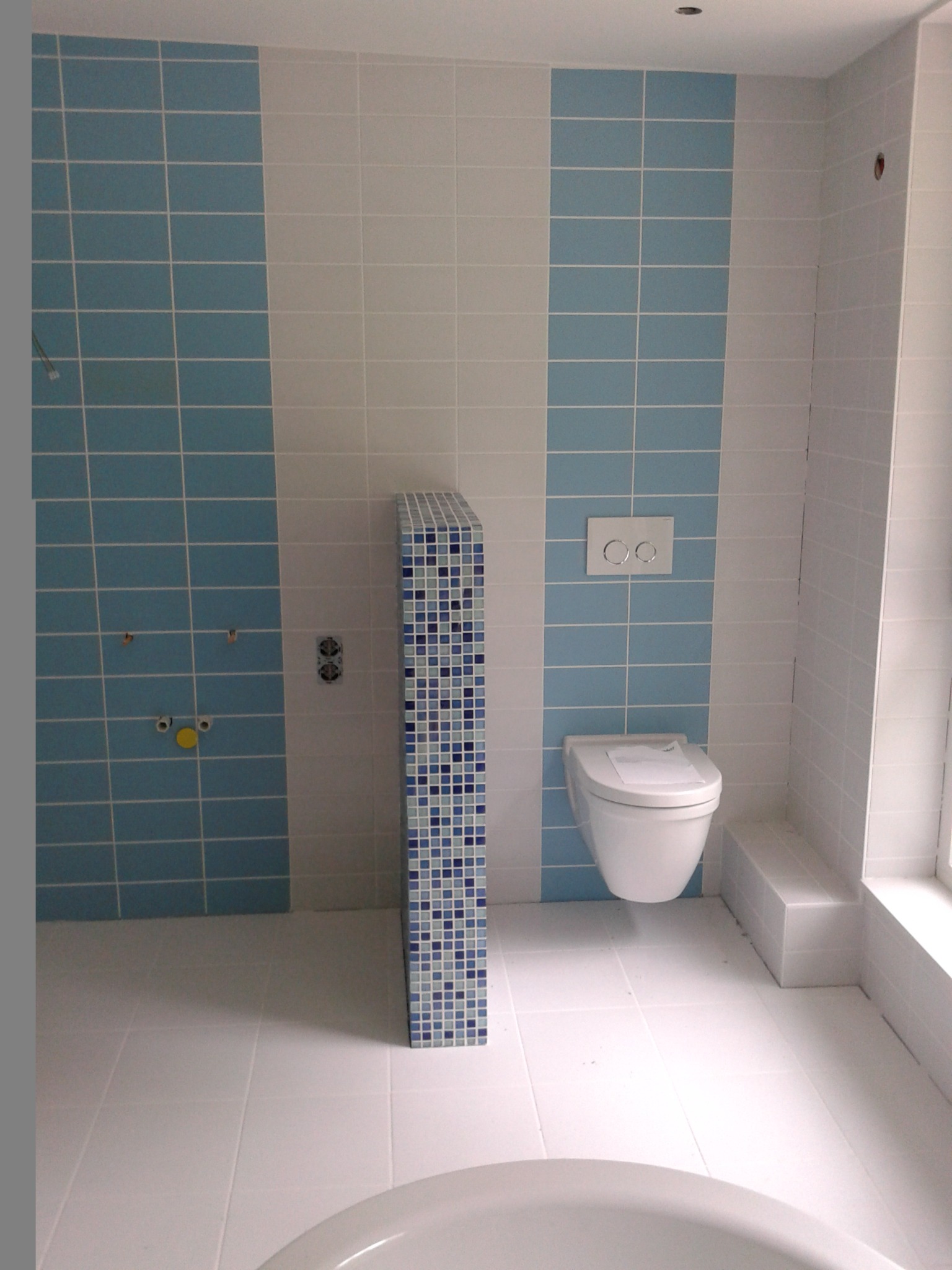 Biało-błękitna łazienka w trakcie remontu, z częściowo ułożonymi płytkami i przygotowanymi instalacjami, widoczna toaleta i pionowa ścianka z mozaiki.