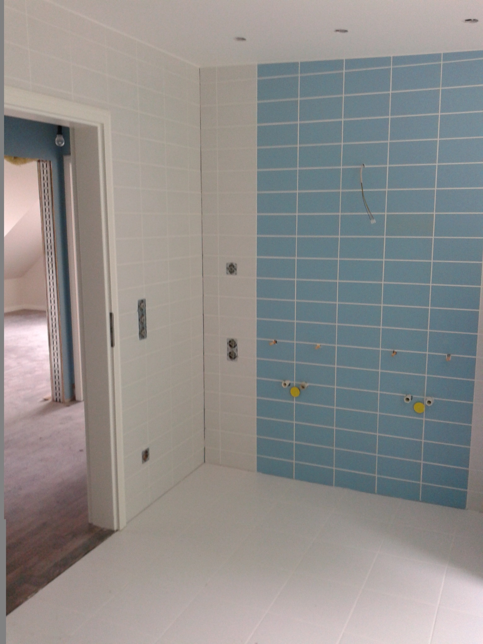 Niedokończona łazienka z białymi i błękitnymi kwadratowymi płytkami, widoczne wyprowadzenia instalacji wodnej i elektrycznej, surowe wykończenie podłogi i fragment sąsiedniego pomieszczenia.