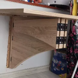 Składany blat drewniany zamontowany na ścianie, widoczne zawiasy i elementy mocujące, w tle suszarka na pranie i inne przedmioty.