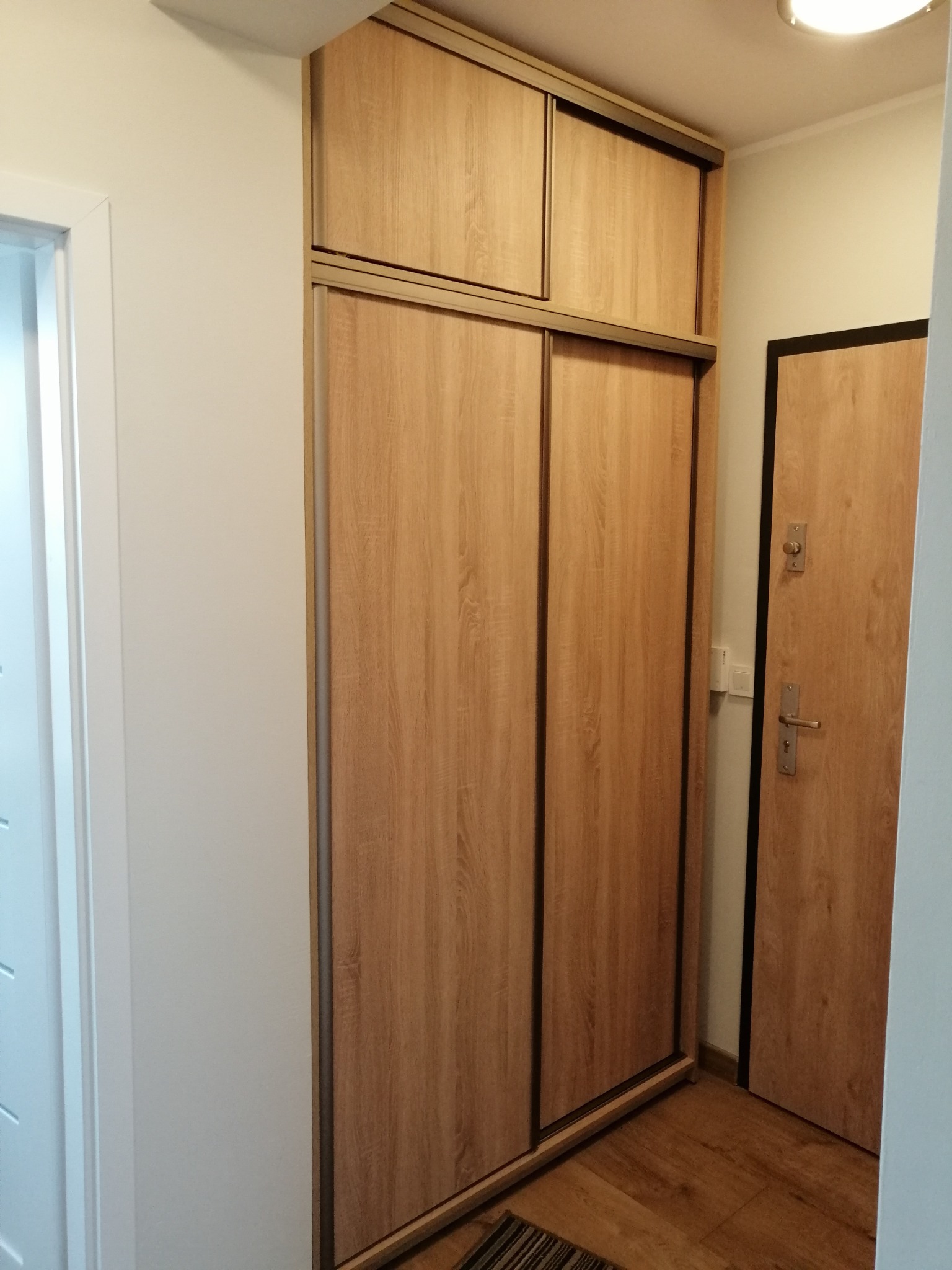 Wysoka szafa przesuwna z drewnopodobnymi frontami w przedpokoju, z dodatkową górną szafką, obok drzwi wejściowych.