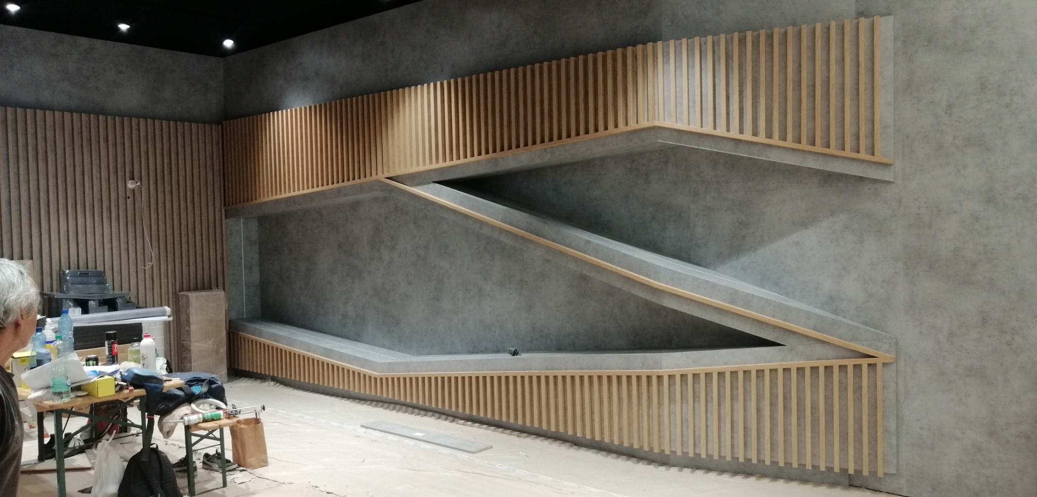 Szara ściana z drewnianymi lamelami i geometrycznym wzorem półek w trakcie montażu, widoczne narzędzia i materiały budowlane na pierwszym planie.