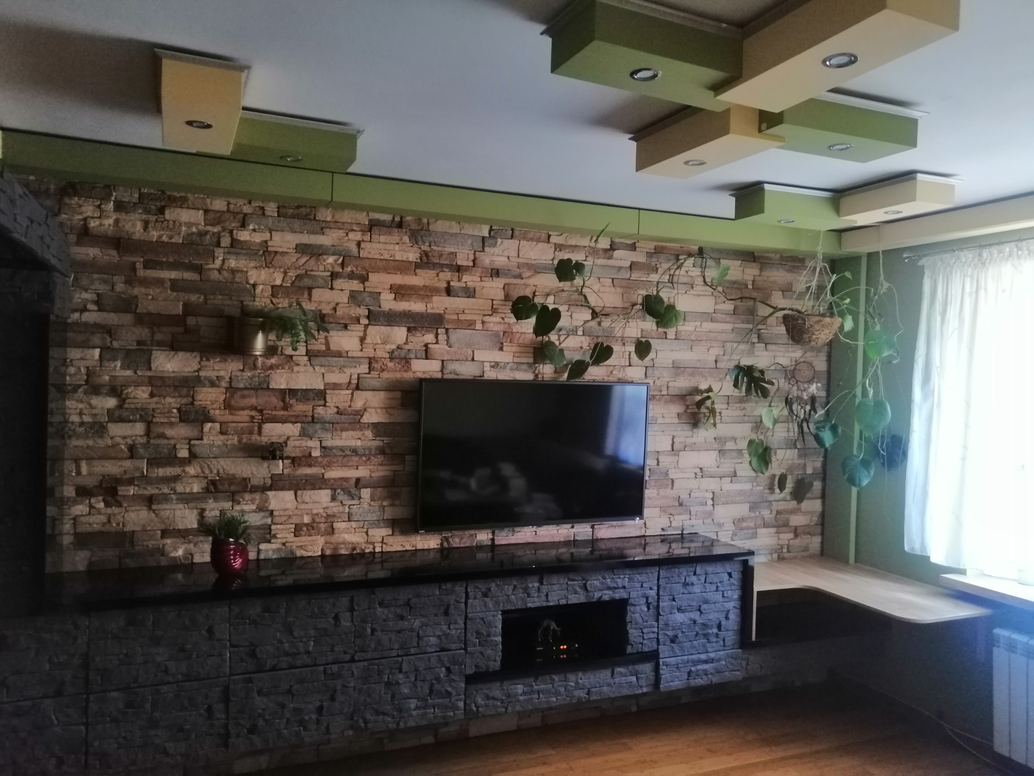 Salon z telewizorem na ścianie z kamienia dekoracyjnego, pod nim szafka RTV z imitacji kamienia, na suficie podwieszany sufit z oświetleniem punktowym, obok okna biurko.