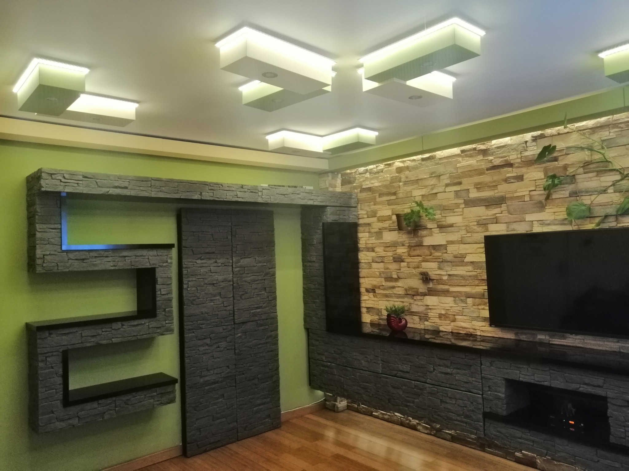 Nowoczesny salon z dekoracyjną ścianą z kamienia, podświetlanymi modułowymi lampami sufitowymi i półkami z efektem kamienia, na podłodze panele drewnopodobne.