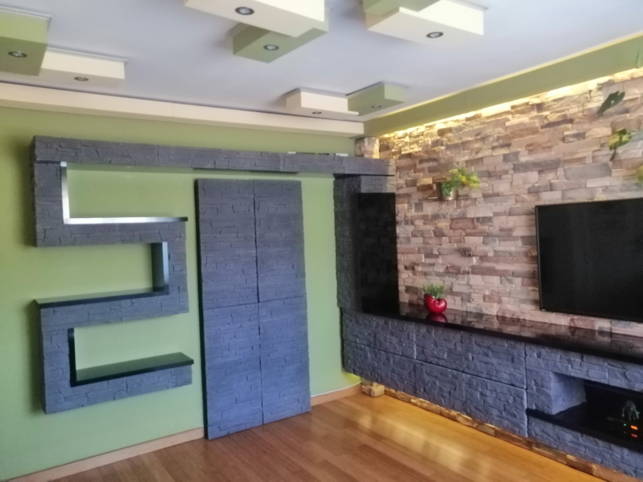 Nowoczesny salon z kamienną ścianą, przesuwanymi drzwiami i półkami z imitacji kamienia, oświetlenie LED pod szafką RTV.