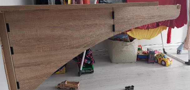 Fragment szafy z jasnego drewna z widocznymi zawiasami, na podłodze zabawki dziecięce i suszarka z praniem w tle.