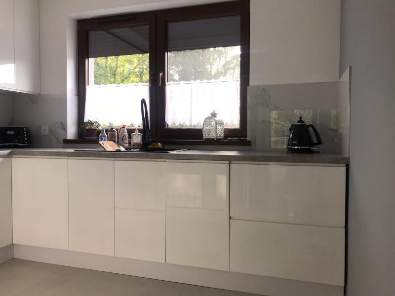 Biała, nowoczesna zabudowa kuchenna z błyszczącymi frontami, szarym blatem i marmurowym panelem ściennym, widok na okno z zasłonką i czarną armaturą.