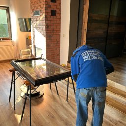 Podlaskiserwisstolarki - Pracownik w niebieskiej bluzie z logo firmy pochylony nad ramą okienną na kozłach w jasnym wnętrzu domu z ceglanym kominem.