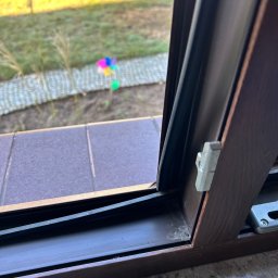 Podlaskiserwisstolarki - Brązowe okno PCV z widocznym uszczelnieniem i mechanizmem blokującym, na tle ogrodu z wiatraczkiem.