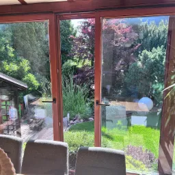 Drewniane okna przesuwne z widokiem na zadbany ogród z oczkiem wodnym i dekoracyjną kulą, widoczne z wnętrza przeszklonej werandy.