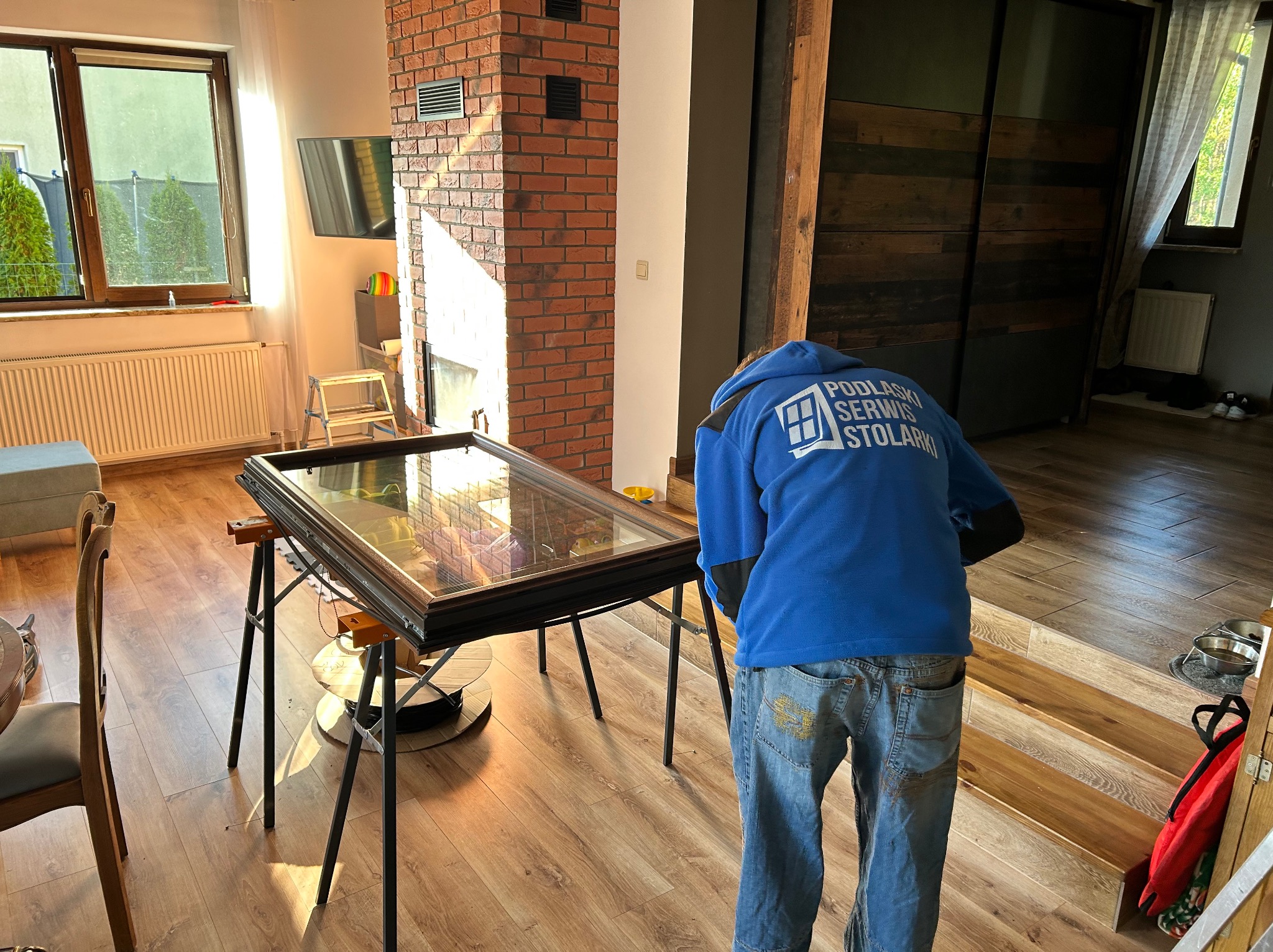 Pracownik w niebieskiej bluzie z logo firmy pochylony nad ramą okienną na kozłach w jasnym wnętrzu domu z ceglanym kominem.