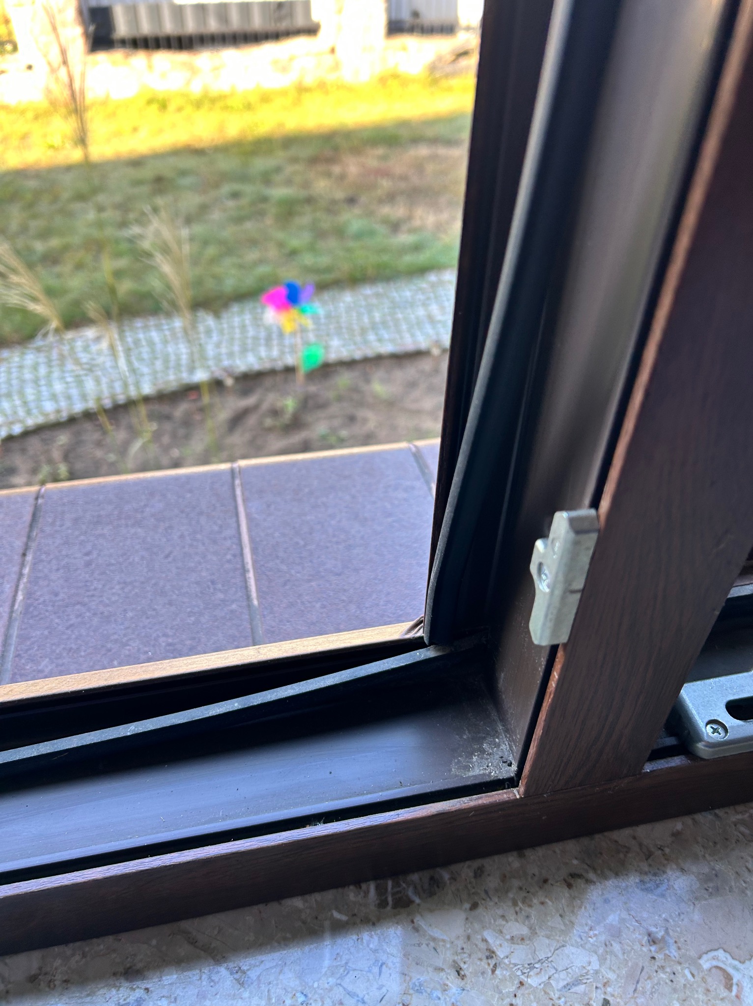 Brązowe okno PCV z widocznym uszczelnieniem i mechanizmem blokującym, na tle ogrodu z wiatraczkiem.